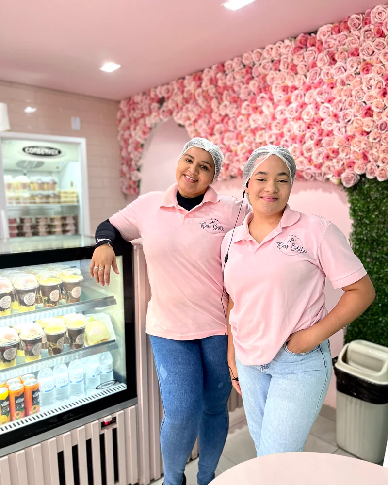 Duas atendentes de uniforme rosa na loja, ao lado da vitrine de doces